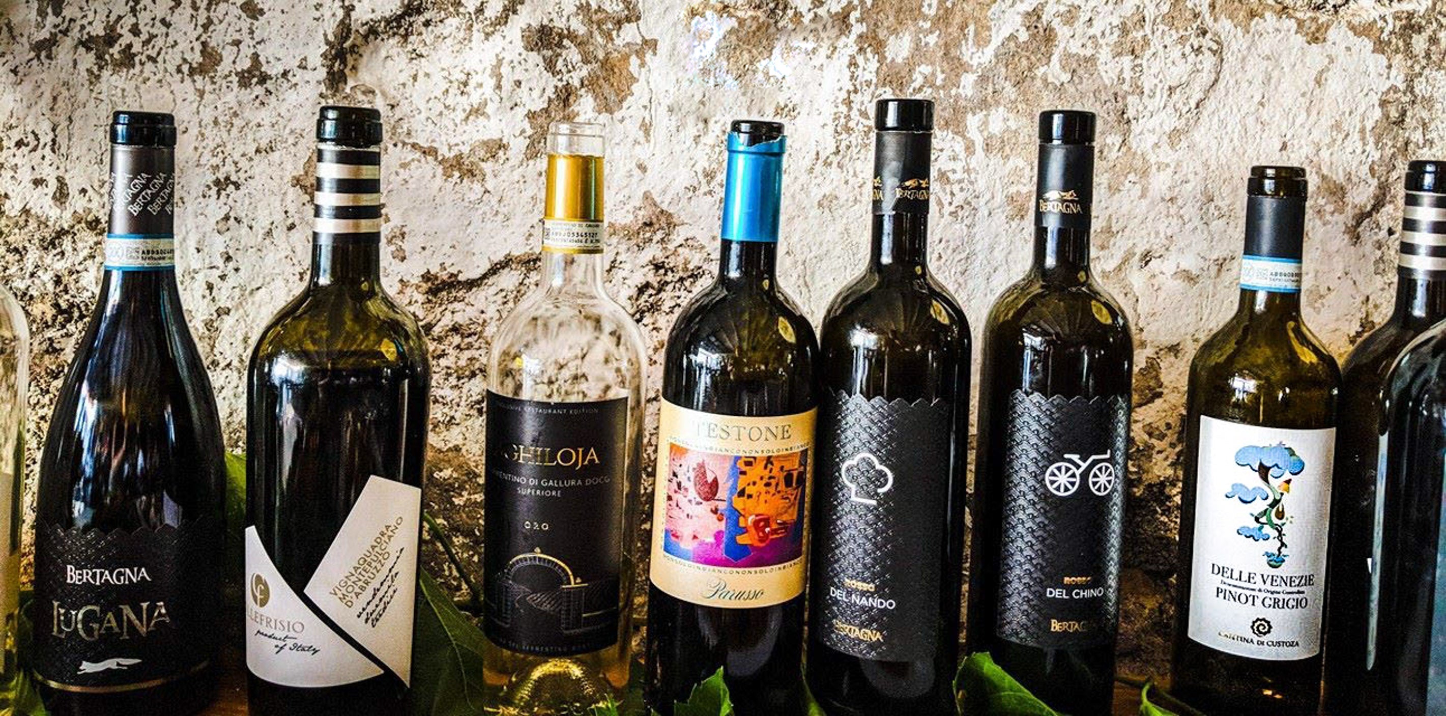 Italienische Weine in Flaschen vor rustikaler Steinwand, Auswahl verschiedener Weinsorten