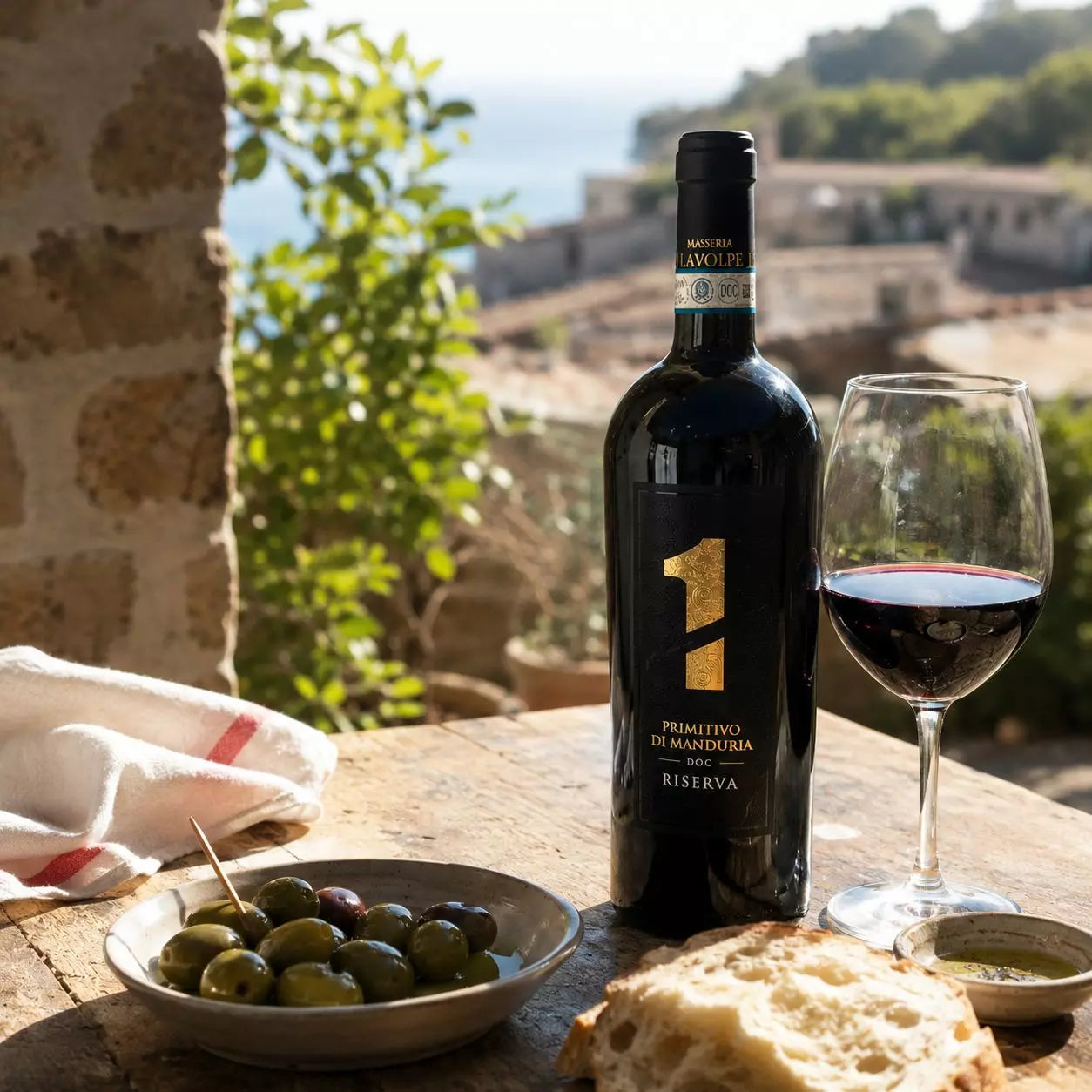 Masseria LaVolpe Primitivo di Manduria Riserva in einer stimmungsvollen italienischen Urlaubsszene auf einer sonnigen Terrasse mit Rotweinglas, Oliven und Brot.